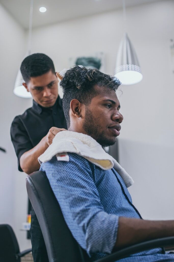 Homem de pele negra sentado em um cabelereiro. O corte de cabelo americano é mais longo no centro da cabeça. O que permite penteados para diversas ocasiões. Na nuca o fade é bem curto. 