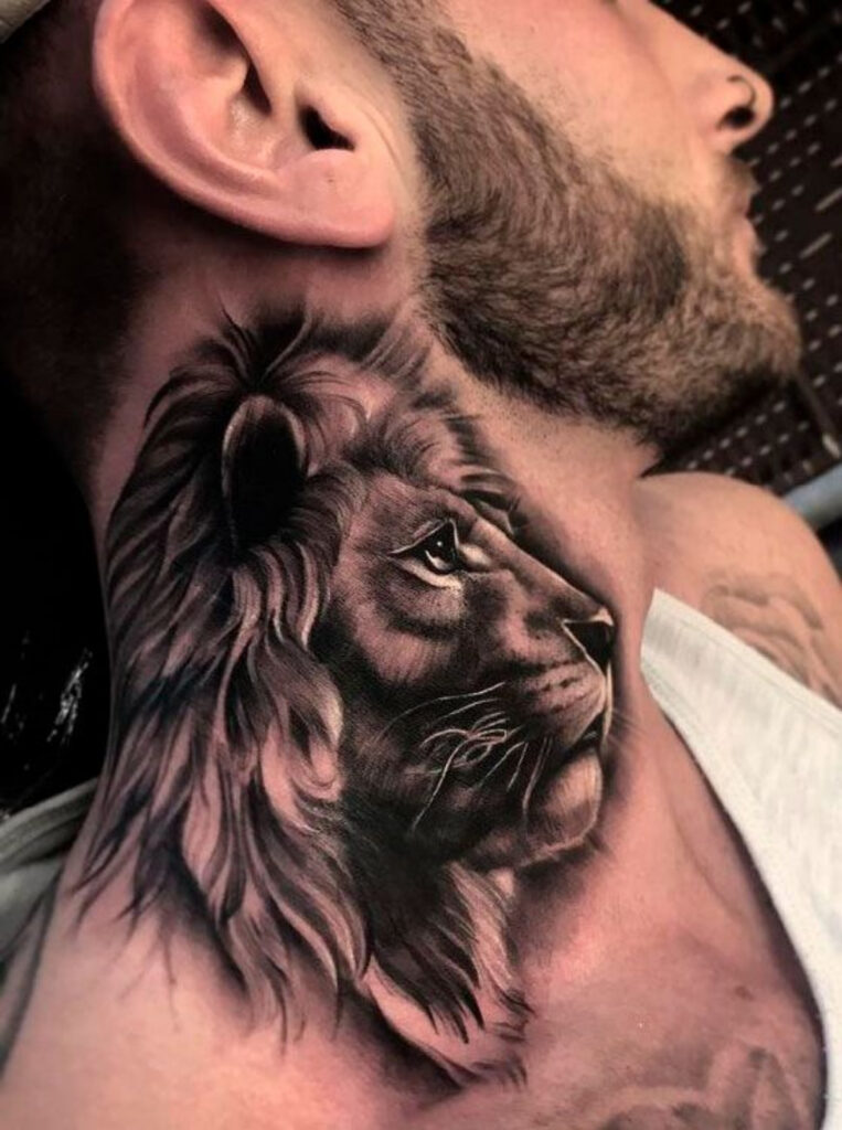 Lateral do pescoço de um homem de pele branca que usa barba, toda tatuada com o rosto de um leão. A imagem está até a juba e mostra o leão imponente olhando para frente, passando a ideia de p roteção. 