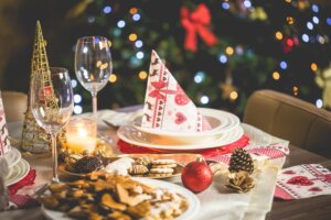 Jantar de natal: mesa de natal com prato com guardanapo natalino em cima da mesma, com taças encima da mesa, bolinhas e pinhas de decoração e comida sobre a mesa, e ao fundo uma árvore-de-natal