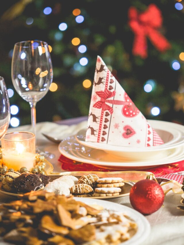 Jantar de natal: mesa de natal com prato com guardanapo natalino em cima da mesma, com taças encima da mesa, bolinhas e pinhas de decoração e comida sobre a mesa, e ao fundo uma árvore-de-natal