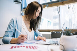 Planejamento financeiro: jovem moça com um casaco azul e uma blusa branca, segurando uma caneta na mão esquerda, desenhando gráficos nas folhas que estão em cima da mesa e com notebook aberto com a mão esquerda no touch pad, o fundo da imagem é um escritório