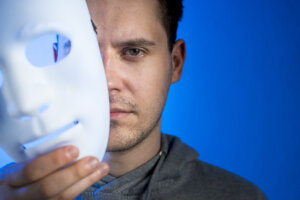 sindrome do impostor: homen segurando uma mascara branca ao lado do rsto estilo hacker com fundo azul
