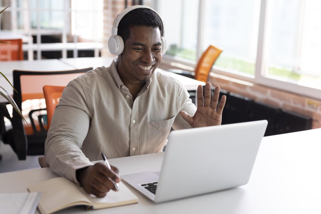 Homem negro acenando para um laptop. Ele sorri e usa um fone de ouvido